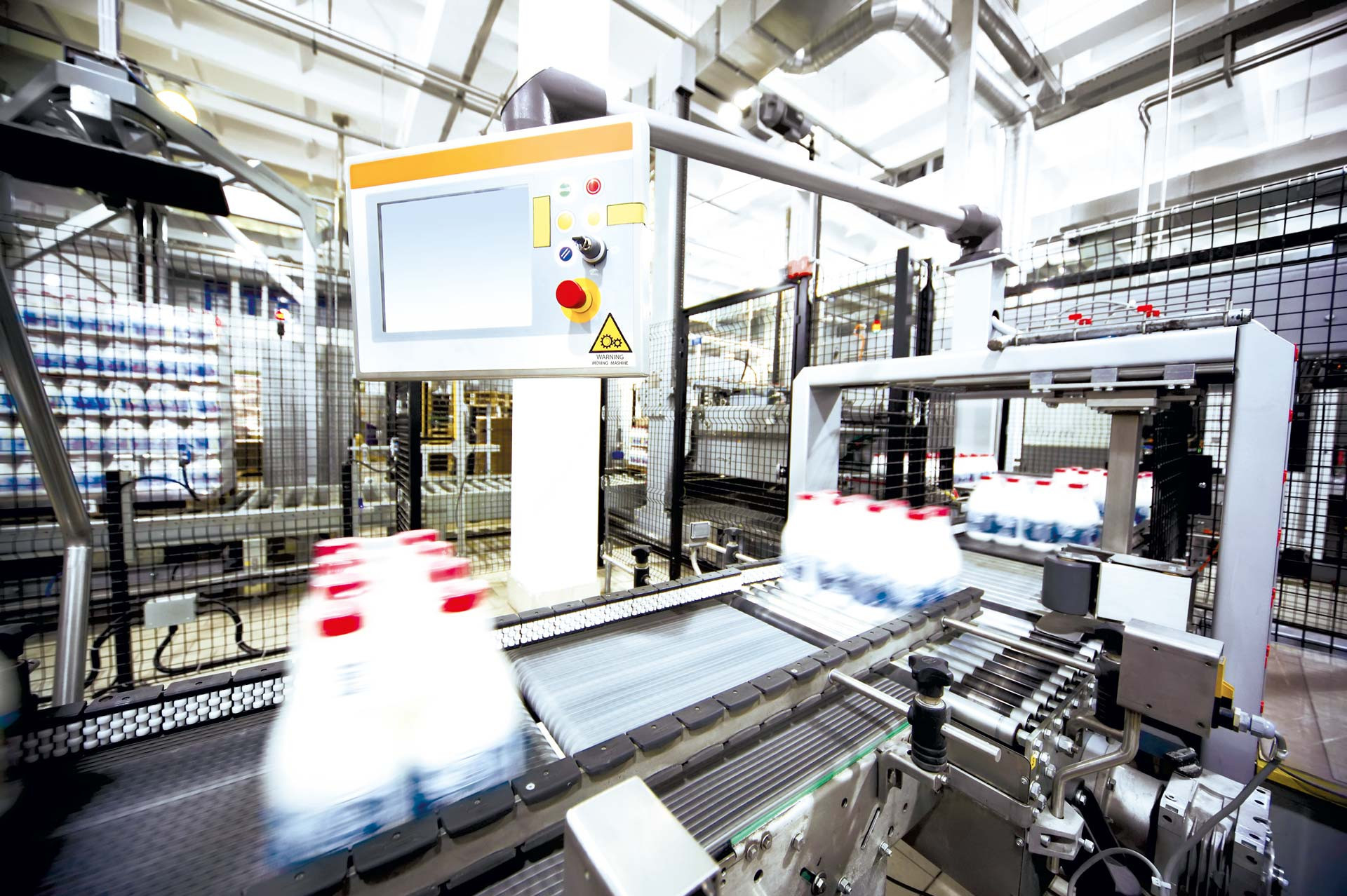 Automatisierte Förderanlage mit Getränkegebinden, gesteuert über Bedienpanel, hinter Schutzgittern in Fabrikhalle.